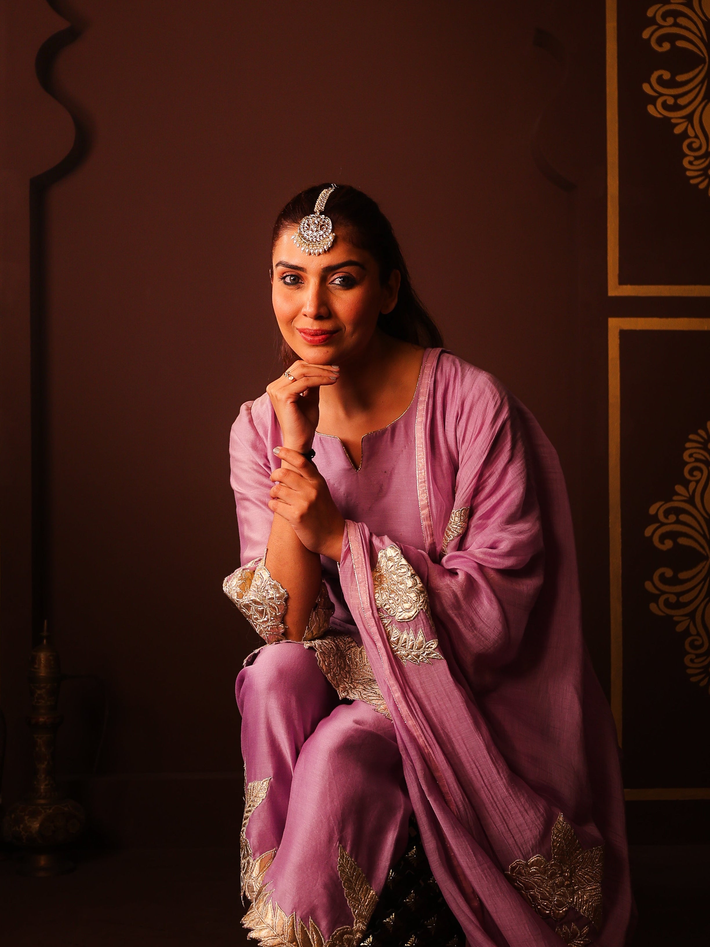 Woman in a traditional pink outfit with gold details sitting against an ornate wall.